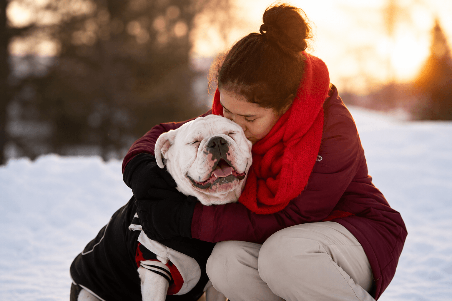 Kristine crouched in the snow at sunset with her arms around Coco, a white American Bulldog in a black coat, her face resting against the dog's head.